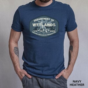 May include: Navy heather t-shirt featuring a Department of Wetlands design. The design includes the text "Department of Wetlands" and "Keeping Resources Productive" with illustrations of birds and plants. The shirt is a short-sleeved crew neck.