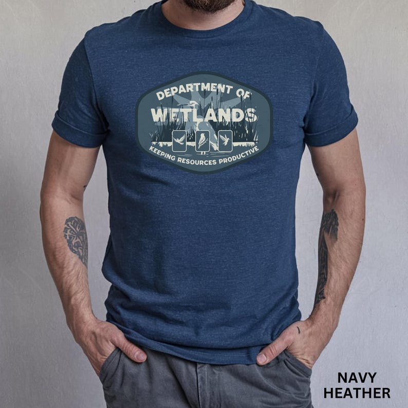 May include: Navy heather t-shirt featuring a Department of Wetlands design. The design includes the text "Department of Wetlands" and "Keeping Resources Productive" with illustrations of birds and plants. The shirt is a short-sleeved crew neck.