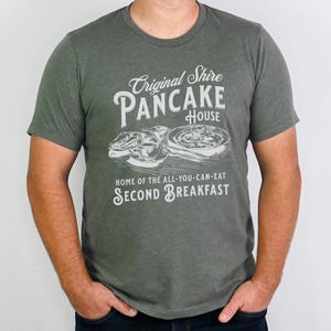 Op de afbeelding: Een groen T-shirt met een wit grafisch ontwerp met de tekst "Original Shire Pancake House" en "Home of the all-you-can-eat Second Breakfast".