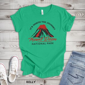 May include: A green t-shirt with the text "It's Worth the Journey" and "Mount Doom National Park" in red and black. The design features a volcano and trees. White sneakers and blue jeans are visible.
