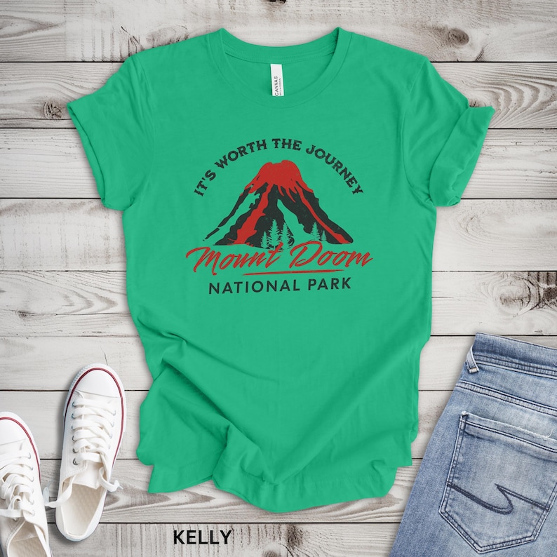 May include: A green t-shirt with the text "It's Worth the Journey" and "Mount Doom National Park" in red and black. The design features a volcano and trees. White sneakers and blue jeans are visible.