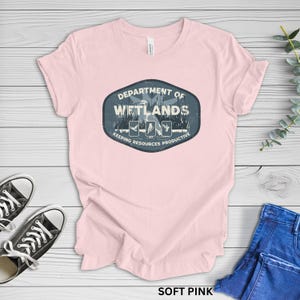 May include: Soft pink t-shirt with a crew neck. The shirt features a graphic on the front that reads "DEPARTMENT OF WETLANDS" in a blue oval, with illustrations of birds and the text "KEEPING RESOURCES PRODUCTIVE". Black sneakers and blue jeans are visible.