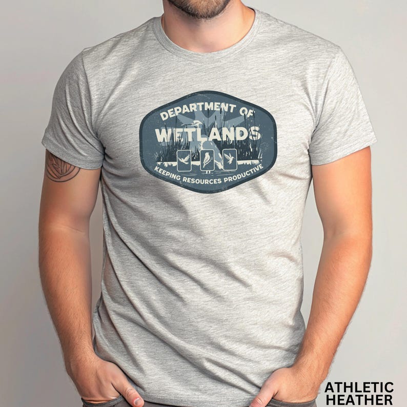 May include: Athletic heather gray t-shirt featuring a blue and white graphic. The graphic reads "DEPARTMENT OF WETLANDS" with illustrations of birds and the text "KEEPING RESOURCES PRODUCTIVE". The shirt is made of a soft, comfortable material.