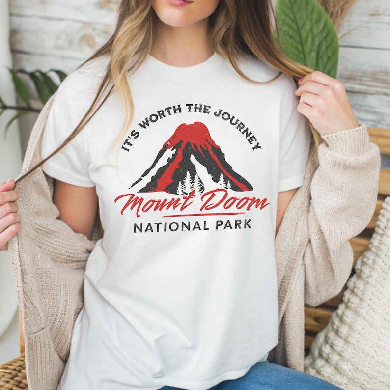 May include: White t-shirt featuring a graphic of a red volcano with the text "It's Worth The Journey" and "Mount Doom National Park". The design includes a mountain and trees. The shirt is a classic crew neck.