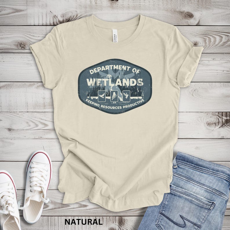 May include: A natural-colored t-shirt with a blue and white graphic that reads "Department of Wetlands" with illustrations of birds and the text "Keeping Resources Productive." The shirt is paired with blue jeans and white sneakers.