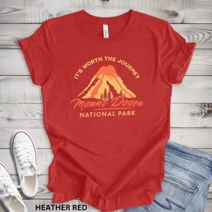 May include: Heather red t-shirt with a graphic of a volcano and the text "It's Worth the Journey Mount Doom National Park". The shirt is paired with white sneakers and blue jeans.