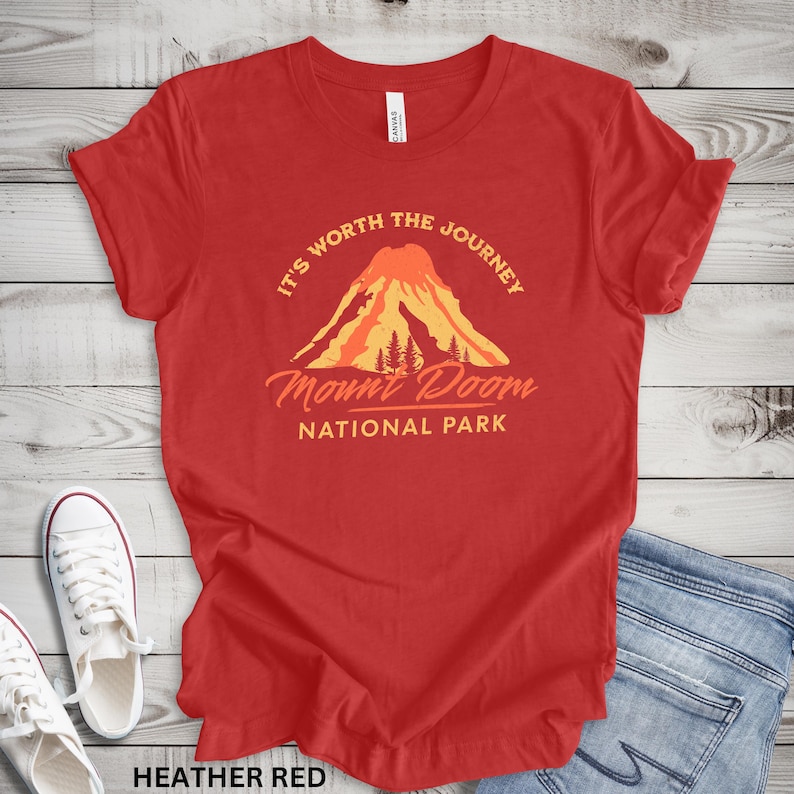 May include: Heather red t-shirt with a graphic of a volcano and the text "It's Worth the Journey Mount Doom National Park". The shirt is paired with white sneakers and blue jeans.