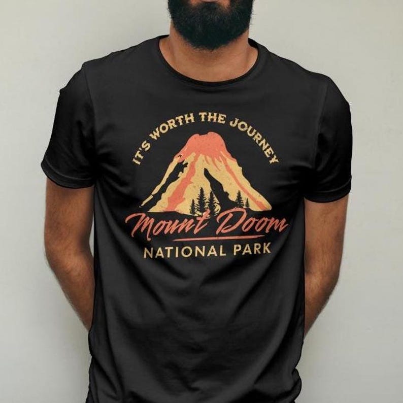 May include: Black t-shirt featuring a mountain graphic with the text "It's Worth the Journey" and "Mount Doom National Park" in a vintage style. The graphic includes a volcano and trees in shades of orange and yellow.
