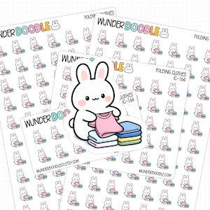 Pode incluir: Várias folhas de adesivos com um coelho de desenho animado dobrando roupas. Os adesivos exibem o texto "WUNDER DOODLE" e "FOLDING CLOTHES IC-164". O coelho segura uma camiseta rosa sobre uma pilha de roupas dobradas.