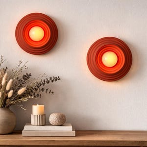 May include: Two terracotta-colored wall sconces with a concentric design and a warm, glowing light. The sconces are mounted on a neutral-colored wall above a wooden table. A vase of dried flowers and a candle are on the table.