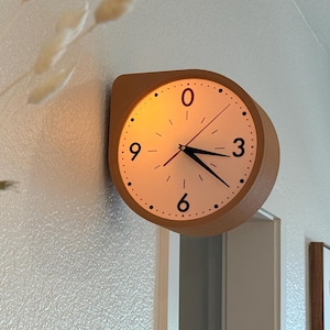 May include: A wall-mounted clock with a light wood frame and a circular face featuring black numerals at the zero, three, six, and nine o'clock positions, along with black hour, minute, and second hands, mounted on a white wall near a doorway.