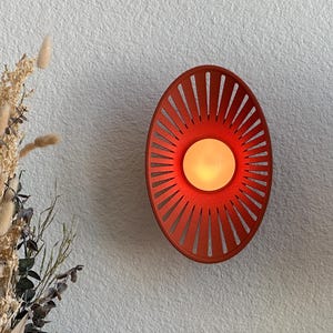 May include: A wall-mounted light fixture featuring a circular, orange, sunburst-style frame with radiating slats. A central, glowing, warm-toned light bulb is positioned in the middle of the frame, casting a soft illumination against the wall.