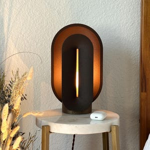May include: A table lamp featuring a dark brown, oval-shaped frame with a recessed, lighter brown inner section. A vertical, glowing light strip is centered within the dark brown core. The lamp sits on a round, white marble-topped side table.