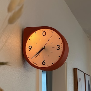 May include: A wall-mounted clock with a terracotta-colored circular frame and a white face. The clock features black numerals at the 0, 3, 6, and 9 positions, black tick marks for the remaining hours, and thin black hour, minute, and second hands.
