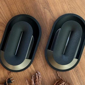 May include: Two modern, black wall sconces with a unique, curved design. Each sconce features a gold-colored accent at the base and a dark gray, textured surface. The sconces are oval-shaped and sit on a wooden surface.