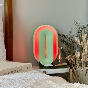 May include: A green and red oval-shaped lamp with a white base. The lamp is on a white nightstand with a book titled "The Kinfolk Home" on top. There are dried flowers in a vase next to the lamp.
