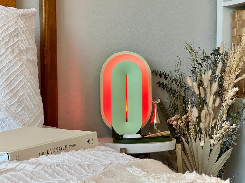May include: A green and red oval-shaped lamp with a white base. The lamp is on a white nightstand with a book titled "The Kinfolk Home" on top. There are dried flowers in a vase next to the lamp.