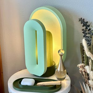 May include: A green and yellow ceramic lamp with a unique geometric design. The lamp is on a white nightstand with a green tray and a small white object.