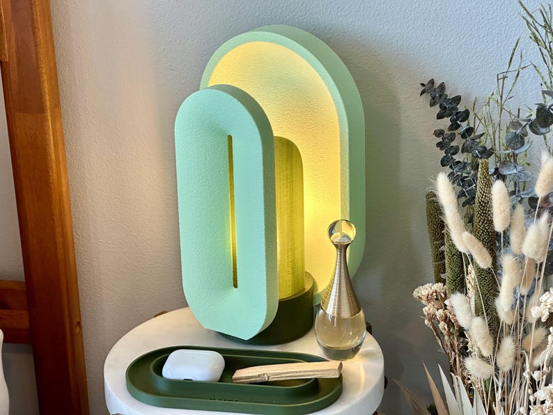 May include: A green and yellow ceramic lamp with a unique geometric design. The lamp is on a white nightstand with a green tray and a small white object.