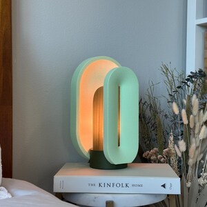 May include: A modern, minimalist table lamp with two overlapping, oval-shaped shades in a light green color. The lamp is lit, casting a warm glow. The lamp is sitting on a book titled "The Kinfolk Home".
