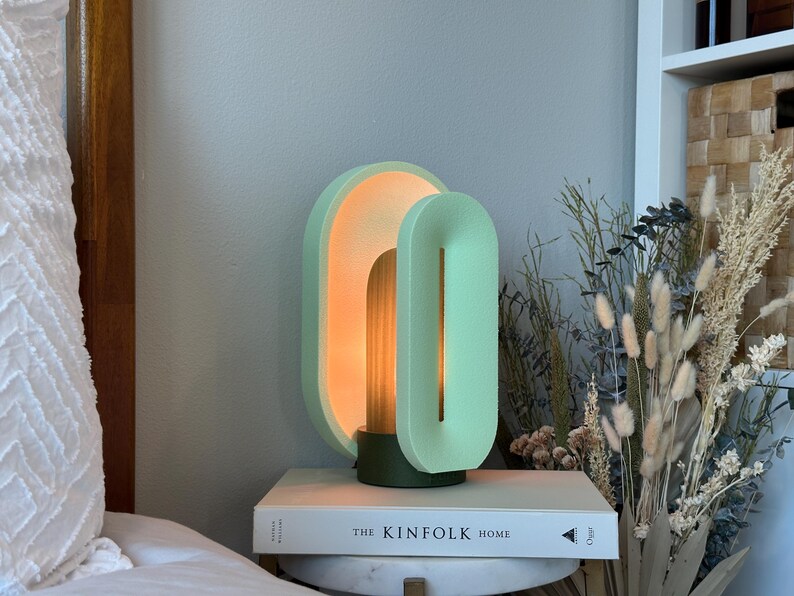 May include: A modern, minimalist table lamp with two overlapping, oval-shaped shades in a light green color. The lamp is lit, casting a warm glow. The lamp is sitting on a book titled "The Kinfolk Home".