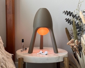 May include: A taupe-colored, three-legged lamp with a textured, matte finish and a rounded top. The lamp features a central, glowing orange spherical light source suspended between its legs. The lamp has a structured, tripod-like base design.