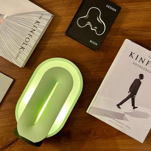 May include: A green oval-shaped lamp with a light inside. The lamp is on a wooden surface with books and magazines around it.
