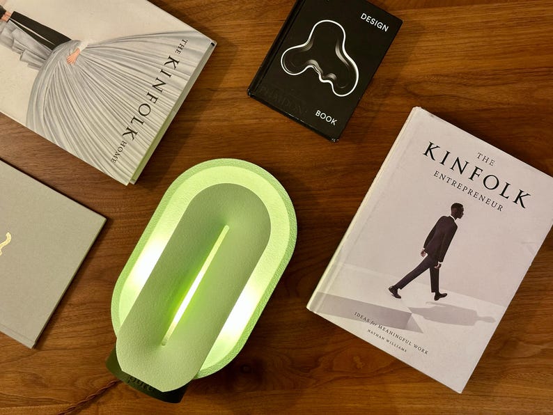 May include: A green oval-shaped lamp with a light inside. The lamp is on a wooden surface with books and magazines around it.