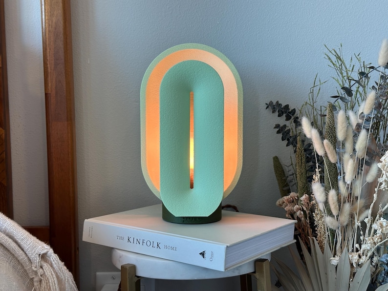 May include: A light green and orange ceramic table lamp with a unique design. The lamp is on a white book with the text "The Kinfolk Home".