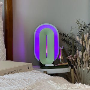 May include: A white and green oval-shaped lamp with a purple light on a bedside table. The lamp is next to a clear glass bottle with a gold cap and a bouquet of dried flowers.