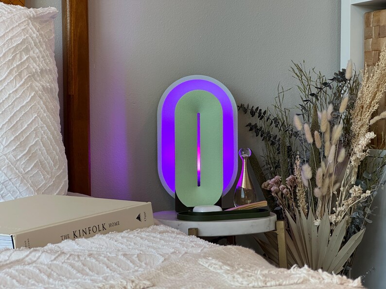 May include: A white and green oval-shaped lamp with a purple light on a bedside table. The lamp is next to a clear glass bottle with a gold cap and a bouquet of dried flowers.