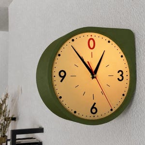May include: A round wall clock with a matte olive green frame and a cream-colored face. The clock features black numerals at the 3, 6, and 9 positions, a red zero at the top, black hour and minute hands, and a thin red second hand.