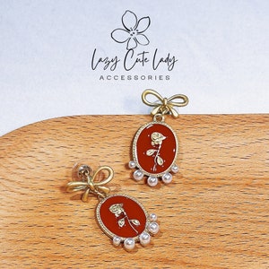 May include: A pair of gold-tone earrings with a red oval-shaped pendant featuring a gold rose design. The pendant is surrounded by a row of white pearl-like beads. The earrings have a bow-shaped top.
