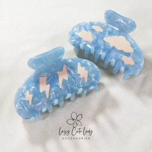 Op de afbeelding: Twee lichtblauwe haarclips met een gemarmerd effect. Eén clip heeft roze bliksemontwerpen, de andere heeft witte wolkvormen. De clips zijn gemaakt van glanzend materiaal en zijn ontworpen om haar op zijn plaats te houden.