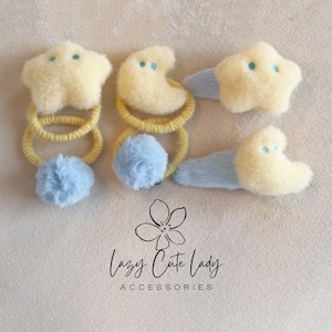 Op de afbeelding: Een verzameling haaraccessoires met hemelse ontwerpen. De set bevat gele haarclips in de vorm van sterren en halve manen en haarbanden met gele banden en blauwe pompons. De tekst "Lazy Cute Lady ACCESSORIES" is ook zichtbaar.
