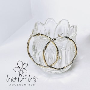 May include: A pair of gold hoop earrings with a textured finish. The earrings are displayed in a clear glass bowl.