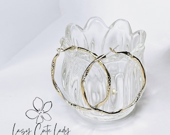 May include: A pair of gold hoop earrings with a textured finish. The earrings are displayed in a clear glass bowl.