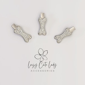 May include: Three silver-toned dog bone-shaped hair clips, each encrusted with sparkling rhinestones. The clips are arranged on a white background. The text "Lazy Cute Lady ACCESSORIES" is below the clips.