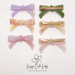 Pu&ograve; includere: Sei fermagli per capelli a forma di fiocco colorati in viola, verde, rosa e pesca. Ogni fermaglio presenta una barretta color oro. Il testo "Lazy Cute Lady Accessories" &egrave; in basso.
