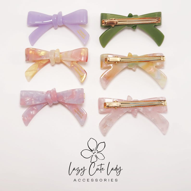 Pu&ograve; includere: Sei fermagli per capelli a forma di fiocco colorati in viola, verde, rosa e pesca. Ogni fermaglio presenta una barretta color oro. Il testo "Lazy Cute Lady Accessories" &egrave; in basso.