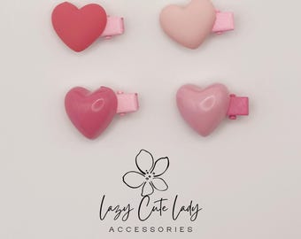 Puede incluir: Cuatro pinzas para el pelo en forma de corazón en tonos de rosa, desde rosa intenso hasta rosa pálido, se muestran sobre un fondo blanco. Cada pinza tiene una base rosa a juego. El texto "Lazy Cute Lady ACCESSORIES" está debajo.