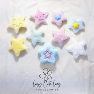 May include: Nine plush star-shaped hair clips in a variety of pastel colours, including pink, blue, yellow, and white. Each star features a smaller star in the centre with a different colour and design. The stars are displayed on a white surface with the text "Lazy Cute Lady ACCESSORIES" at the bottom.