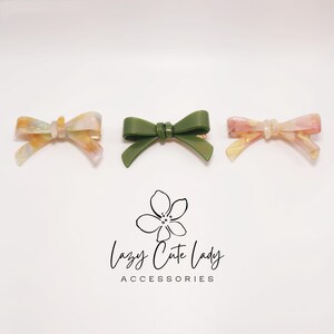 Pu&ograve; includere: Tre fermagli per capelli a forma di fiocco. Due fermagli presentano un motivo marmorizzato nei toni del giallo, rosa e bianco. Il terzo fermaglio &egrave; verde oliva. I fermagli sono su sfondo bianco. Il testo "Lazy Cute Lady ACCESSORIES" &egrave; sotto.