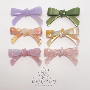 May include: Six colorful bow hair clips in shades of purple, green, pink, and yellow. Each clip has a bow shape with a textured finish. The text "FRANCE" is visible on some of the bows. The image also includes the text "Lazy Cute Lady Accessories".