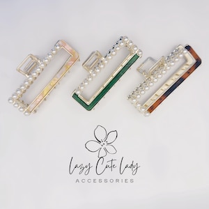 Puede incluir: Tres pinzas para el pelo rectangulares. Una es dorada con un diseño veteado rosa y amarillo, otra es verde y la tercera es carey. Cada pinza está delineada con pequeñas perlas blancas de imitación. La marca "Lazy Cute Lady Accessories" está en la parte inferior.