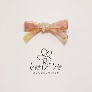 Pu&ograve; includere: Fermaglio per capelli a forma di fiocco nei toni del rosa, giallo e bianco. La clip ha un effetto marmorizzato e una chiusura a barretta. Il testo "Lazy Cute Lady ACCESSORIES" &egrave; sotto.