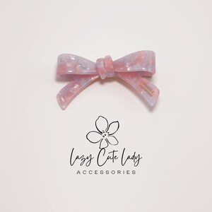 Pu&ograve; includere: Fermaglio per capelli a forma di fiocco marmorizzato rosa chiaro e lilla. Il fiocco &egrave; realizzato in materiale lucido e riporta la scritta "FRANCE Handmade" su una delle code. Il logo "Lazy Cute Lady Accessories" &egrave; sotto.