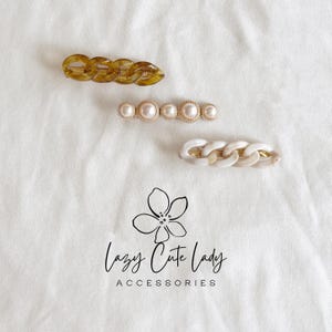Könnte beinhalten: Drei Haarspangen auf weißem Hintergrund. Eine hat ein Kettenglied-Design in Schildpattfarbe. Eine andere hat Kunstperlen. Die dritte ist ein weißes Kettenglied-Design. Das Logo "Lazy Cute Lady Accessories" befindet sich unten.