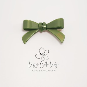 Pu&ograve; includere: Un fermaglio per capelli a forma di fiocco verde con la scritta "FRANCE Handmade" su un lato. Il fermaglio &egrave; realizzato in materiale lucido ed &egrave; posto su uno sfondo bianco. L'immagine include anche il testo "Lazy Cute Lady ACCESSORIES".