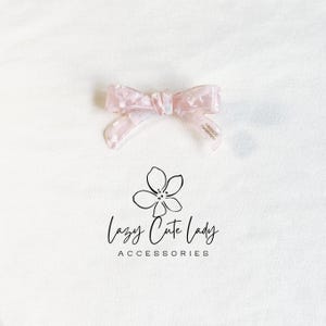 Pu&ograve; includere: Un fiocco rosa chiaro per capelli con un motivo marmorizzato. Il fiocco &egrave; realizzato in un materiale traslucido e presenta la scritta "FRANCE Handmade" su una delle code. Il logo "Lazy Cute Lady Accessories" &egrave; sotto il fiocco.